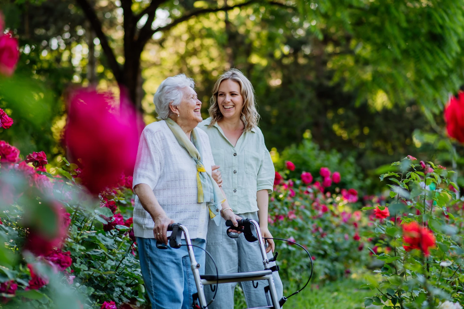 Junge Frau begleitet ältere Frau mit Rollator durch einen blühenden Rosengarten. Beide lachen und wirken gelöst.
