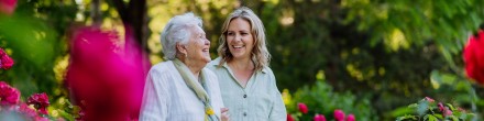 Junge Frau begleitet ältere Frau mit Rollator durch einen blühenden Rosengarten. Beide lachen und wirken gelöst.