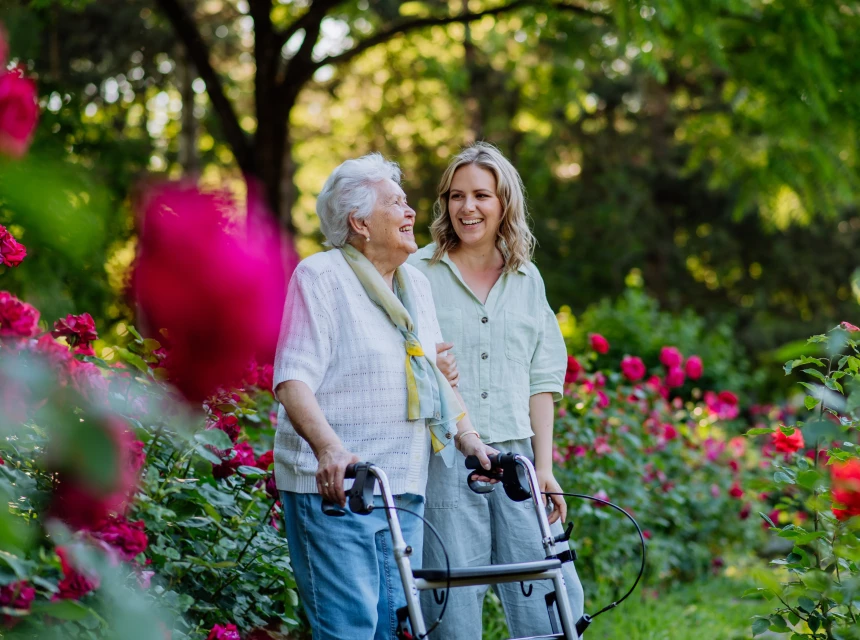 Junge Frau begleitet ältere Frau mit Rollator durch einen blühenden Rosengarten. Beide lachen und wirken gelöst.