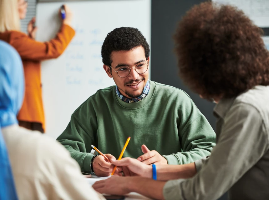 Gruppe junger Erwachsener diskutiert in einem Kursraum, ein Mann mit Brille spricht, andere hören zu und schreiben mit.